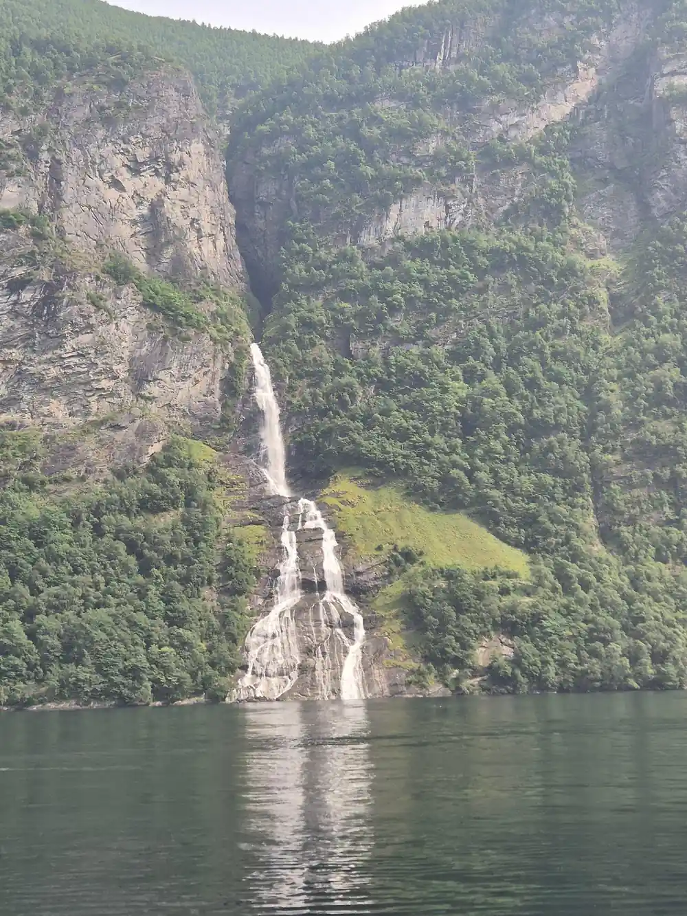 Tall slender waterfall on steep mountain cliff above fjord water